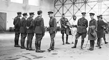 A group of military personnel in an aircraft hangar, four of whom are in a row facing another man, while the remainder stand informally, one of them wearing a flying suit