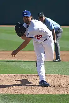 A man wearing a white baseball uniform and a blue baseball cap follows through after throwing a baseball