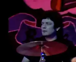 A male drummer is shown in upper shot behind his kit (it is not visible). His head is turned to his right. He wears a dark T-shirt partly obscured by a cymbal on its stand in front of the kit. He also has a jewelled earring visible in his left ear. The backdrop depicts part of the group's logo with the image of a rose partly visible.