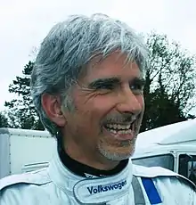 Damon Hill sporting a goatee in white racing overalls smiling to the left of the camera