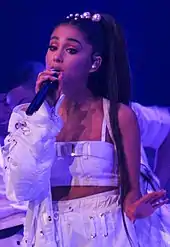 An zoomed-in image of a woman dressed in a white crop top, pants and a jacket performing on stage.