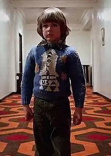 Danny Lloyd as the child character Danny Torrance in the 1980 film The Shining. He stands in a hotel hallway and wears a sweater with a rocketship on it.