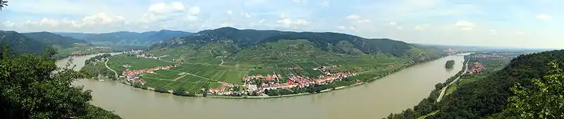 A vast river curves through a flat landscape. At the end of the flood 7plain, steep mountains rise above red-roofed houses.