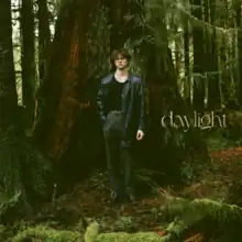 A man in a black top, trousers, and leather jacket standing afront trees in a forest. The lowercase text "daylight" stands on his left.