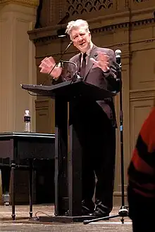 A white-haired man in a black suit stands on a stage behind a black podium; both his hands are raised. A microphone is visible is visible in front of him and a second microphone is seen to the right. A bottle of water is visible on a black table to the left.