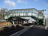 Two people walking on a green bridge with a white lattice running over a railway track and platform under a blue sky with white clouds