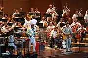 Five members of a band play keyboards, electric bass, drums and guitar in front of a symphony orchestra and its conductor, all dressed informally.