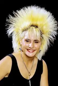 A teenage girl in a black tank top and large, blonde spiky hair smiles to the camera on a black background.