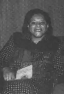 A young African-American woman, seated and smiling, wearing a print dress and holding a pamphlet in her lap.