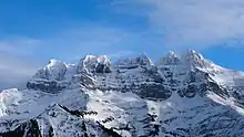 Vertical sawtooth-shaped rock walls overhanging a snowy slope lined with a few trees.