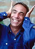 Derek Jarman wearing a denim shirt, smiling and putting hands against his ears