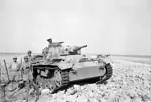 A tank lies crippled in the desert with soldiers standing around it