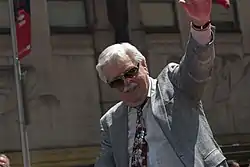 Caucasian man with white hair and mustache raising his left arm. He is wearing sunglasses, a jacket and tie.