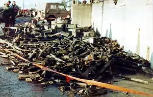 A large pile of military hardware, including rifles, machine guns, small arms ammunition. It is roped off with red plastic tape. Three civilians look on.