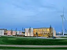 This is the front view of the DkIT's Nursing Building. It is a picture of mutistoreyed glass building with white walls as borders on both sides. There are similar looking buildings on the left. There is a windmill on the right. The building is lit with yellow lights. There are trees planted in front of the building. The lawn in front of the building is neatly manicured. The pathway between the lawn and the building has walkway lights (yellow). The sky above is clear blue.