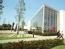 This is the side view of the Nursing Building. It is a glass building with white walls as borders. The picture is taken during the day. There are two or three tall trees in front, with two cone like decorative structures in front. There are two groups of students (groups of 3 and 2 respectively) walking on the pathway. In the background, there are similar looking buildings on the right, and trees on the left. In the foreground, there is a neatly manicured lawn with small hedges.