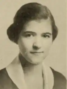 A young white woman with dark hair combed from a sidepart into an updo; she is wearing a dark garment with a white collar