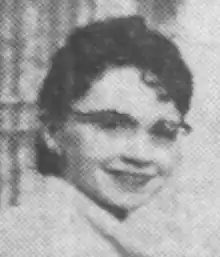 A white woman with short dark curly hair, smiling, wearing eyeglasses.