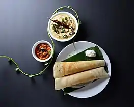 Two dosa rest next to a dollop of butter on a plaintain leaf. There are separate bowls for the sauces.