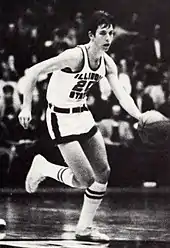 A man, wearing a black suit and a tie, is making a gesture by raising his hands while standing on the side of a basketball court.