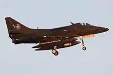 Colour photo of a green military jet aircraft in flight with its landing gear extended