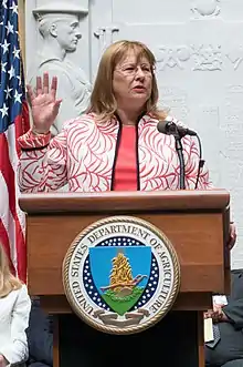 A woman with reddish blonde hair and glasses holds her right hand up as she speaks at a podium with a United States Department of Agriculture seal on it. She wears an embroidered jacket that looks like coral colored plants or leave and a coral shirt underneath.