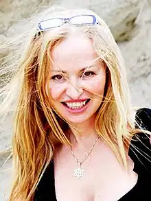 A blonde woman smiling. Around her neck is a necklace containing a religious symbol.