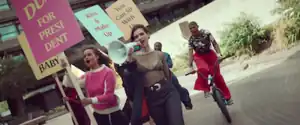 Dua Lipa singing in a megaphone with her girl group who hold rainbow flags and colourful signs with many sayings.