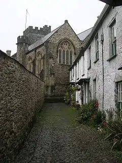 Stone building with arched windows and square tower seen at the end of a narrow lane with white painted houses on the right and a wall on the left.