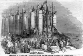 An engraving of a large crowd surrounding a partly complete temple structure, which is topped with flags and missing its entablature