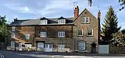 A row of English stone houses with white windows and doors beside a road. There is a stone wall and wooden gate on one side, and several different types of trees around them.