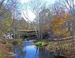 A creek with some waterfalls over an old dam in the distance on a clear autumn day. There is a house on the right obscured by the trees