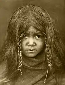 A black-and-white portrait of a young Quilcene boy with long hair and two braids, in the year 1913