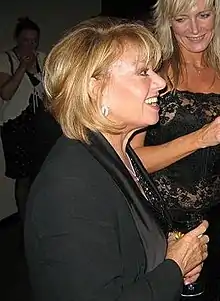 A smiling blonde woman wearing a light blue blouse and an elaborate necklace stares at the camera.