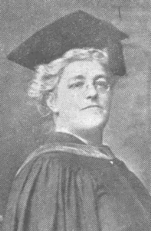 An older white woman in academic regalia, wearing glasses. Her hair is wavy and white under the mortar board on her head.