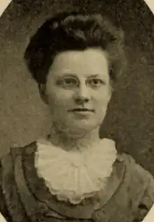 A white woman with dark hair in a bouffant style, wearing glasses and a white ruffle at her neck