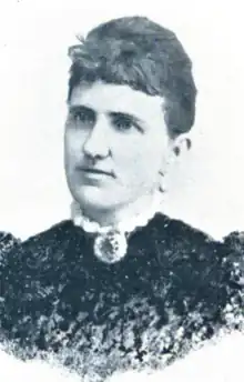 A white woman with dark hair dressed in an updo with short fringe, wearing a dark dress with a high lace-trimmed collar and a brooch fastened at the throat