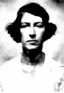 A white woman with dark bobbed hair, parted on the side, wearing a light-colored top with a round neckline