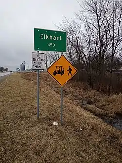 Large green sign in foreground reading "Elkhart, 450", mounted on two metal posts, with a smaller white sign reading "No soliciting without permit" and a yellow diamond-shaped sign with a drawing of a forklift and a person walking; grain elevator in background