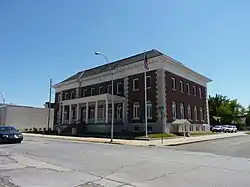 A brick building seen from a corner across the street. It is two stories high with a pointed roof, stone sections at the corners and a white front porch. There is a streetlight and an American flag in front.