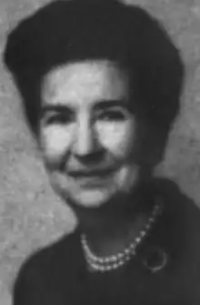 A smiling middle-aged white woman with dark hair in a bouffant style, wearing pearls and a dark suit