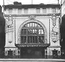 Black and white photo of a theater with a vaulted ceiling and an ornate curtain in front of the stage.