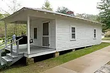 Present-day photograph of a whitewashed house, about 15 feet wide. Four bannistered steps in the foreground lead up to a roofed porch that holds a swing wide enough for two. The front of the house has a door and a single-paned window. The visible side of the house, about 30 feet long, has double-paned windows.