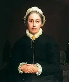 Portrait of woman sitting with hands clasped, wearing a white bonnet and black jacket