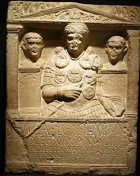 A stone carving, the border resembling a Greco-Roman building, displaying three busts, slightly damaged, the centremost being that of Marcus Caelius, wearing armour, a cape and holding a staff of office in his right hand. Below the carving is a Latin inscription.