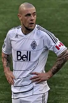 A man wearing a white T-shirt and white shorts, standing on a soccer field with his hands at on his hips, looking to his left.