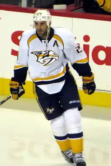 Upper body of a hockey player who is wearing a white uniform with "DALLAS 24" on his chest. He is leaning against another player's stick as he anticipates a faceoff.