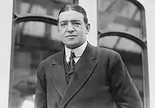  Dark-haired man wearing a tight collar and dark suit, unsmiling, looks directly at camera