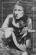 A man dressed as a fool crouches on a straw covered floor with a rough stone wall behind him. He holds a mock scepter in his right hand.