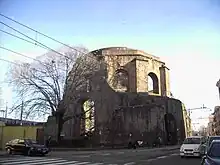 Exterior of a ten-sided ruin called today the Temple of Minerva Medica at the intersection of city streets in Rome showing large arched windows in the drum between engaged buttresses and below polygonal step-rings buttresses for the collapsed dome